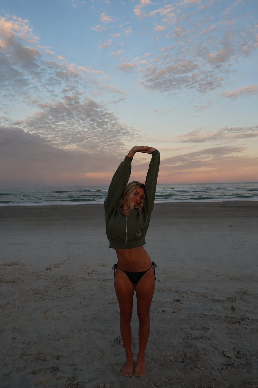 Customer on the beach at sunset wearing the olive Lotus Original cropped zip-up hoodie.